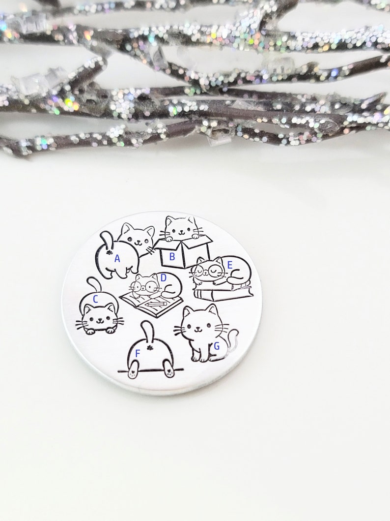 May include: A silver charm with a black and white illustration of seven cats in various poses. The cats are labeled with letters A through G.