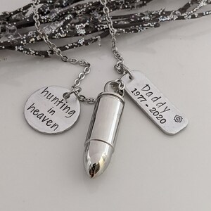 May include: A silver-toned necklace featuring a bullet-shaped pendant, a round charm with "hunting in heaven" text, and a rectangular tag with "Daddy 1977-2020" text. The necklace is displayed against a sparkly, silver-toned branch.