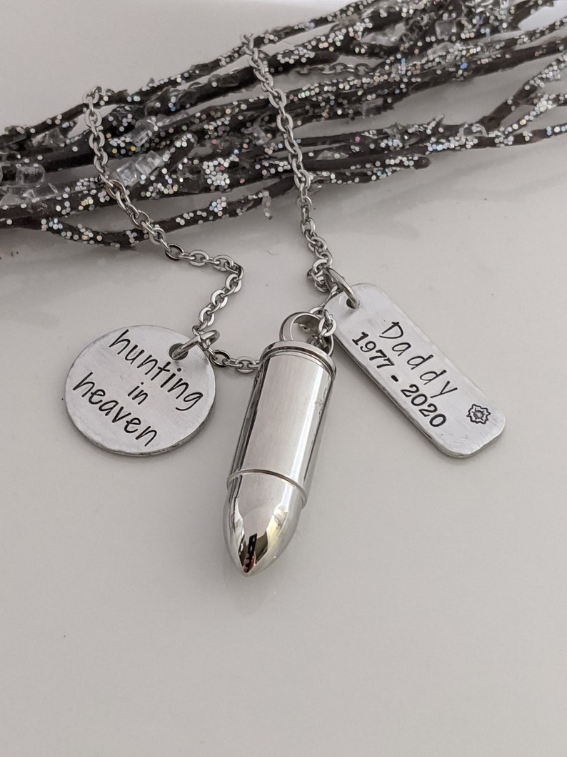 May include: A silver-toned necklace featuring a bullet-shaped pendant, a round charm with "hunting in heaven" text, and a rectangular tag with "Daddy 1977-2020" text. The necklace is displayed against a sparkly, silver-toned branch.