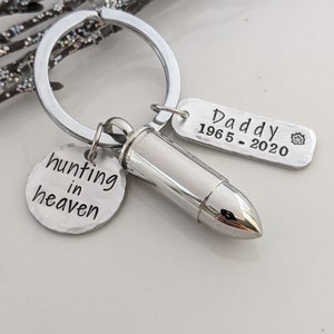 May include: A silver-colored keychain with a bullet-shaped charm, a round charm with the words "hunting in heaven," and a rectangular charm with "Daddy 1965-2020" engraved. The keychain is designed to hold keys.