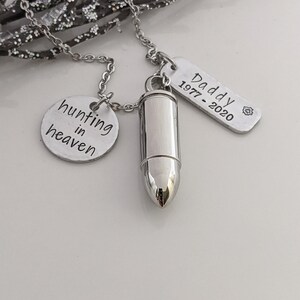 May include: A silver-toned necklace with three pendants. One pendant is a bullet-shaped urn, another is a round charm with the words "hunting in heaven", and the third is a rectangular tag that reads "Daddy 1977-2020".