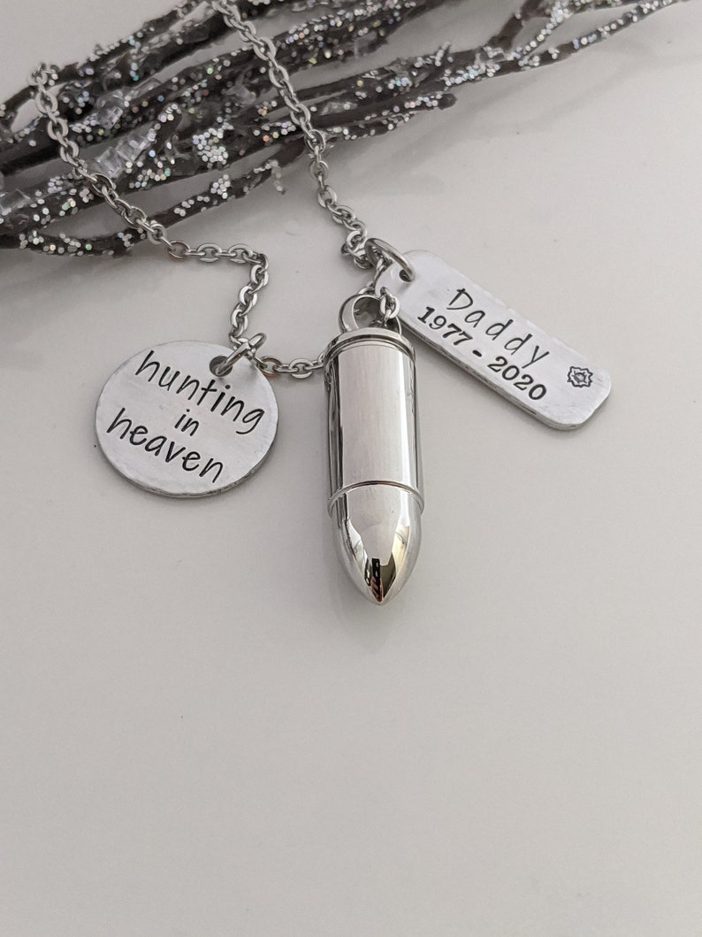 May include: A silver-toned necklace with three pendants. One pendant is a bullet-shaped urn, another is a round charm with the words "hunting in heaven", and the third is a rectangular tag that reads "Daddy 1977-2020".
