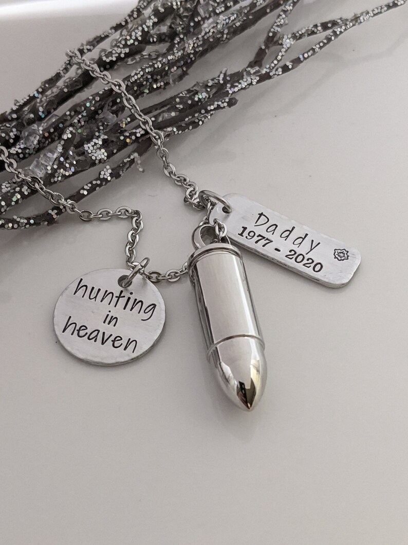 May include: Silver-toned memorial necklace with a bullet-shaped pendant, a round charm with "hunting in heaven" text, and a rectangular tag with "Daddy 1977-2020" engraved. The necklace is displayed on a white surface with glittery branches.