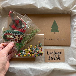 May include: A clear plastic bag filled with Christmas-themed craft supplies, including ribbon, twine, wooden stars, and colorful confetti. A brown card with a green Christmas tree and the words "Christmas Wishes" is also visible, along with a small card that says "Vintage by Sarah".