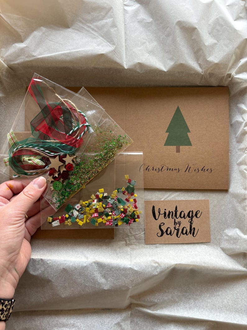 May include: A clear plastic bag filled with Christmas-themed craft supplies, including ribbon, twine, wooden stars, and colorful confetti. A brown card with a green Christmas tree and the words "Christmas Wishes" is also visible, along with a small card that says "Vintage by Sarah".