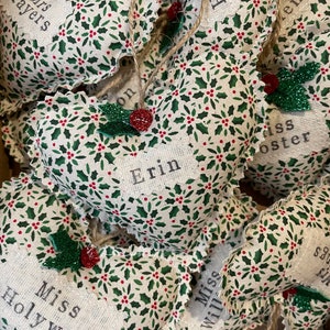 May include: A collection of handmade heart-shaped ornaments with holly and berry patterns. Each ornament has a personalized name, including "Mrs. Mayers", "Erin", "Miss Holywell", and "Miss Will", among others.