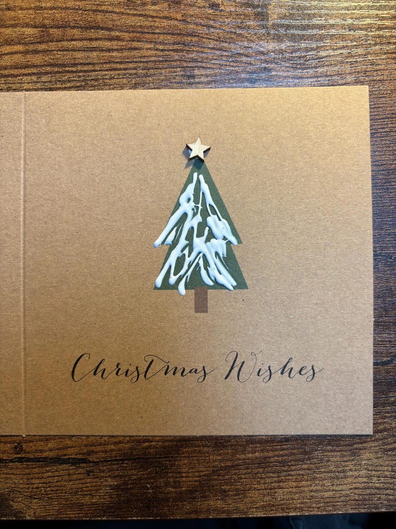 May include: A Christmas card with a green tree design, adorned with white accents and a wooden star. The card is a natural brown color, with the words "Christmas Wishes" written in elegant script.