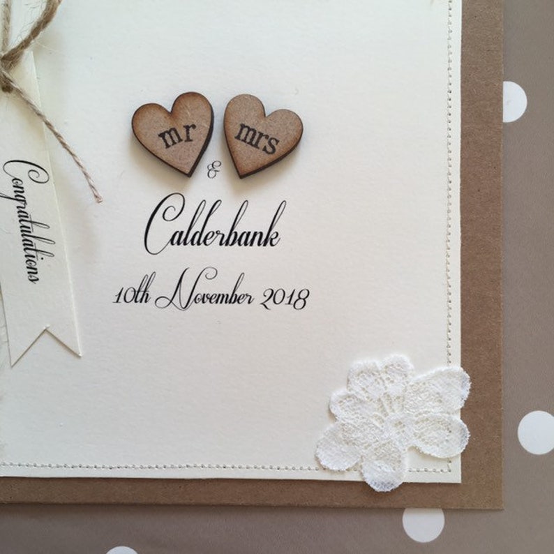 Pu&ograve; includere: Un biglietto bianco con un bordo marrone e un ricamo in pizzo. Il biglietto ha le parole "Mr & Mrs Calderbank" e "10th November 2018" stampate su di esso. Due piccoli cuori di legno con le parole "Mr" e "Mrs" sono attaccati alla parte superiore del biglietto.