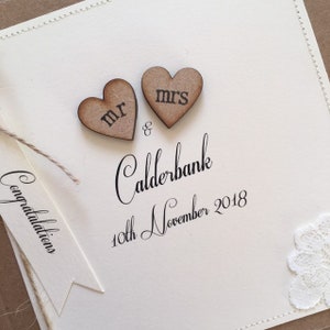 Pu&ograve; includere: Un biglietto bianco con le parole "Congratulations Mr. & Mrs. Calderbank 10th November 2018" scritte in corsivo nero. Due cuori di legno con le parole "Mr." e "Mrs." sono attaccati in cima al biglietto.