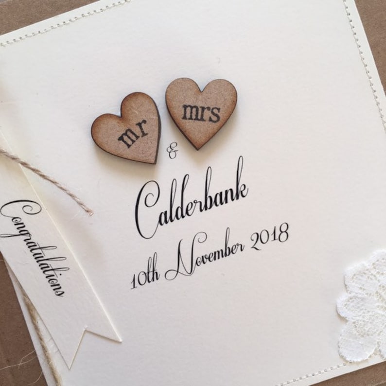 Pu&ograve; includere: Un biglietto bianco con le parole "Congratulations Mr. & Mrs. Calderbank 10th November 2018" scritte in corsivo nero. Due cuori di legno con le parole "Mr." e "Mrs." sono attaccati in cima al biglietto.