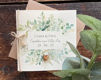 Puede incluir: Una tarjeta blanca con una ilustración de acuarela de hojas verdes y un corazón de madera. La tarjeta dice "Ciara & Paul Felicidades por su día de boda 29.04.23".