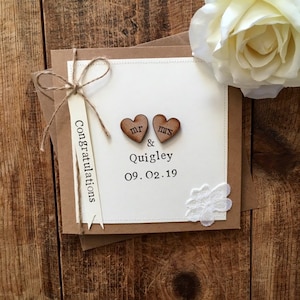 K&ouml;nnte beinhalten: Eine wei&szlig;e Karte mit braunem Rand und einer braunen Kordelschleife. Die Karte hat zwei Holzh Herzen mit den Worten "Mr." und "Mrs." darauf. Die Karte hat auch den Text "Congratulations Mr. & Quigley 09.02.19".