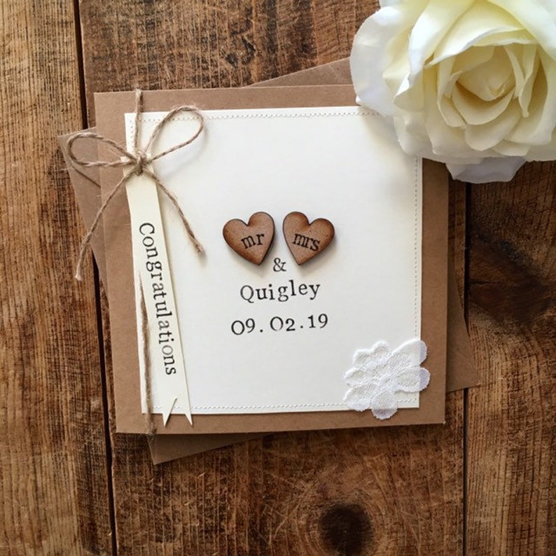 K&ouml;nnte beinhalten: Eine wei&szlig;e Karte mit braunem Rand und einer braunen Kordelschleife. Die Karte hat zwei Holzh Herzen mit den Worten "Mr." und "Mrs." darauf. Die Karte hat auch den Text "Congratulations Mr. & Quigley 09.02.19".