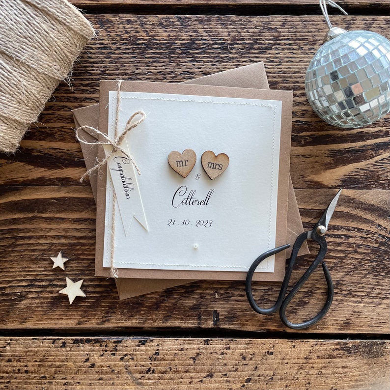 Pu&ograve; includere: Un biglietto bianco con una busta marrone, decorato con due cuori di legno che dicono "Mr" e "Mrs" e il testo "Congratulations Mr & Mrs Cotterell 21.10.2023".