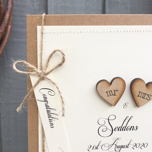 Pu&ograve; includere: Un biglietto di nozze con uno sfondo marrone rustico e un interno di carta bianco. La carta interna ha un'etichetta con la parola "Congratulazioni" scritta su di essa. Due cuori di legno con le parole "Mr." e "Mrs." sono attaccati alla carta. Il testo "Seddon's" e "21 agosto 2020" &egrave; scritto sulla carta.