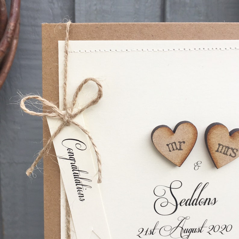 Pu&ograve; includere: Un biglietto di nozze con uno sfondo marrone rustico e un interno di carta bianco. La carta interna ha un'etichetta con la parola "Congratulazioni" scritta su di essa. Due cuori di legno con le parole "Mr." e "Mrs." sono attaccati alla carta. Il testo "Seddon's" e "21 agosto 2020" &egrave; scritto sulla carta.