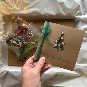 May include: A Christmas card with a decorated tree and the words "Christmas Wishes" is held in a hand. The card is brown with a green ribbon. A package of Christmas decorations and fairy lights are in the background.