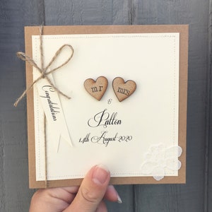 Pu&ograve; includere: Un biglietto di nozze beige con un bordo di carta marrone. Il biglietto ha un fiocco di spago marrone con un'etichetta che dice "Congratulazioni". Due cuori di legno con "Mr." e "Mrs." scritti su di essi sono attaccati al biglietto. Il biglietto ha anche un piccolo pezzo di pizzo bianco e il testo "& Patton 14 agosto 2020".