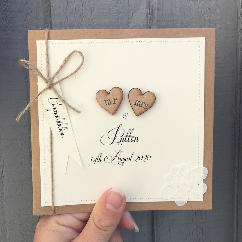 Pu&ograve; includere: Un biglietto di nozze beige con un bordo di carta marrone. Il biglietto ha un fiocco di spago marrone con un'etichetta che dice "Congratulazioni". Due cuori di legno con "Mr." e "Mrs." scritti su di essi sono attaccati al biglietto. Il biglietto ha anche un piccolo pezzo di pizzo bianco e il testo "& Patton 14 agosto 2020".