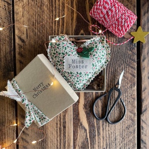 May include: A heart-shaped Christmas ornament with a holly berry pattern, personalized with "Miss Foster," rests in a box. A gift box with "Merry Christmas" printed on it is tied with a ribbon. Twine, scissors, and a star are also present.