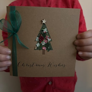 May include: A handmade Christmas card with a brown paper background. The card features a cutout Christmas tree filled with decorative elements and a wooden star on top. The words "Christmas Wishes" are written in cursive, and a green ribbon is tied on the left side.