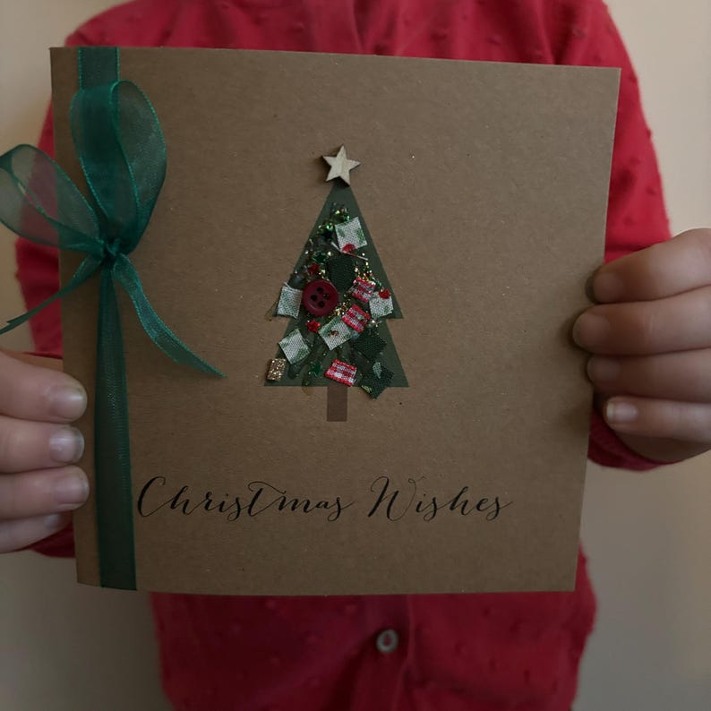 May include: A handmade Christmas card with a brown paper background. The card features a cutout Christmas tree filled with decorative elements and a wooden star on top. The words "Christmas Wishes" are written in cursive, and a green ribbon is tied on the left side.