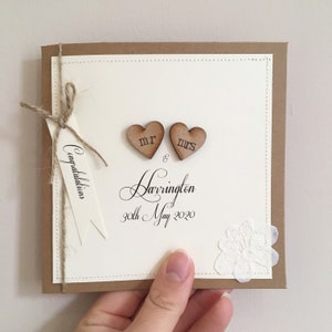 Pu&ograve; includere: Un biglietto bianco con un bordo marrone e un fiocco di spago marrone. Il biglietto ha due cuori di legno con "Mr" e "Mrs" scritti su di essi. Il biglietto ha anche il testo "Congratulations Mr & Mrs Harrington 30th May 2020".