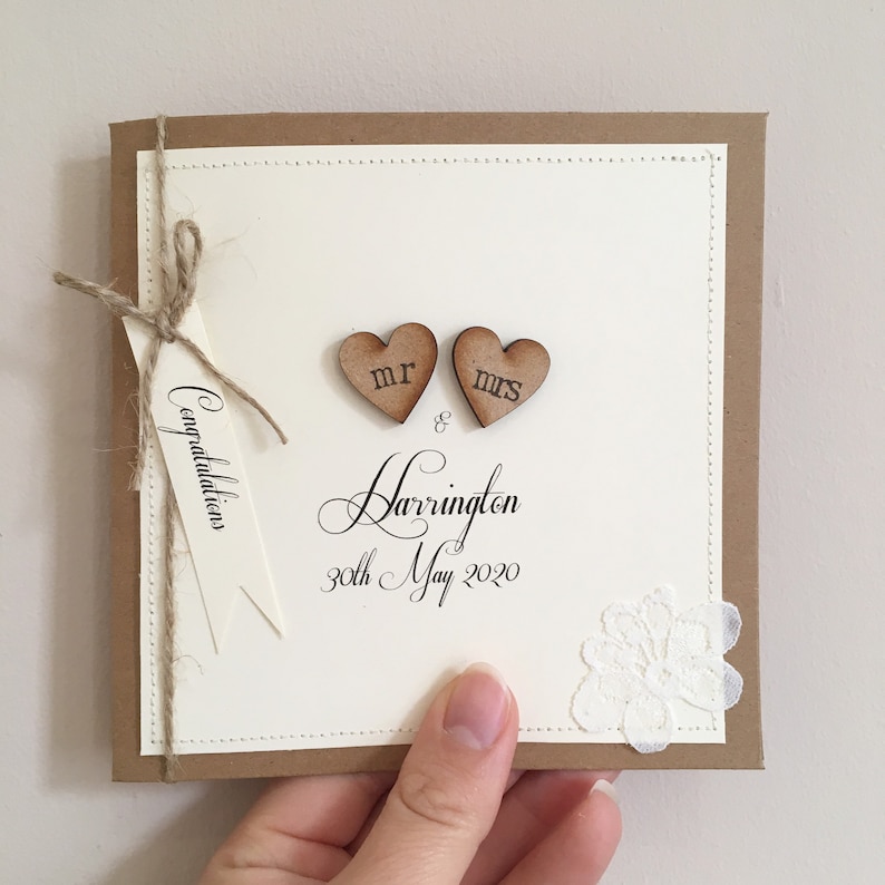 Pu&ograve; includere: Un biglietto bianco con un bordo marrone e un fiocco di spago marrone. Il biglietto ha due cuori di legno con "Mr" e "Mrs" scritti su di essi. Il biglietto ha anche il testo "Congratulations Mr & Mrs Harrington 30th May 2020".