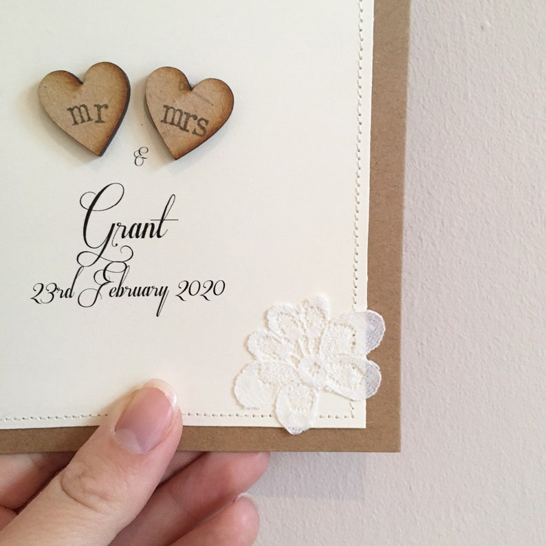 Pu&ograve; includere: Un biglietto bianco con un bordo marrone e un ricamo in pizzo. Il biglietto ha due cuori di legno con le parole "Mr." e "Mrs." su di essi. Il biglietto ha anche il testo "Grant & 23rd February 2020."