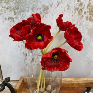 May include: A bouquet of red poppy flowers in a clear glass vase with small stones at the bottom. The vase is sitting on a wooden tray.