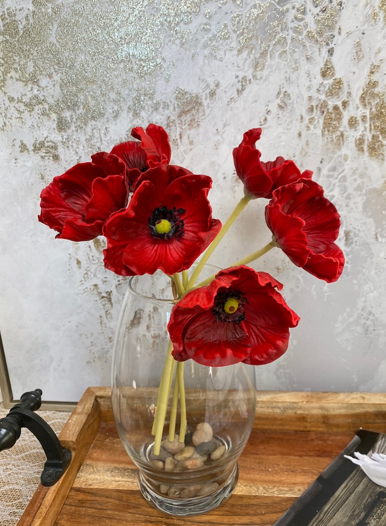 May include: A bouquet of red poppy flowers in a clear glass vase with small stones at the bottom. The vase is sitting on a wooden tray.