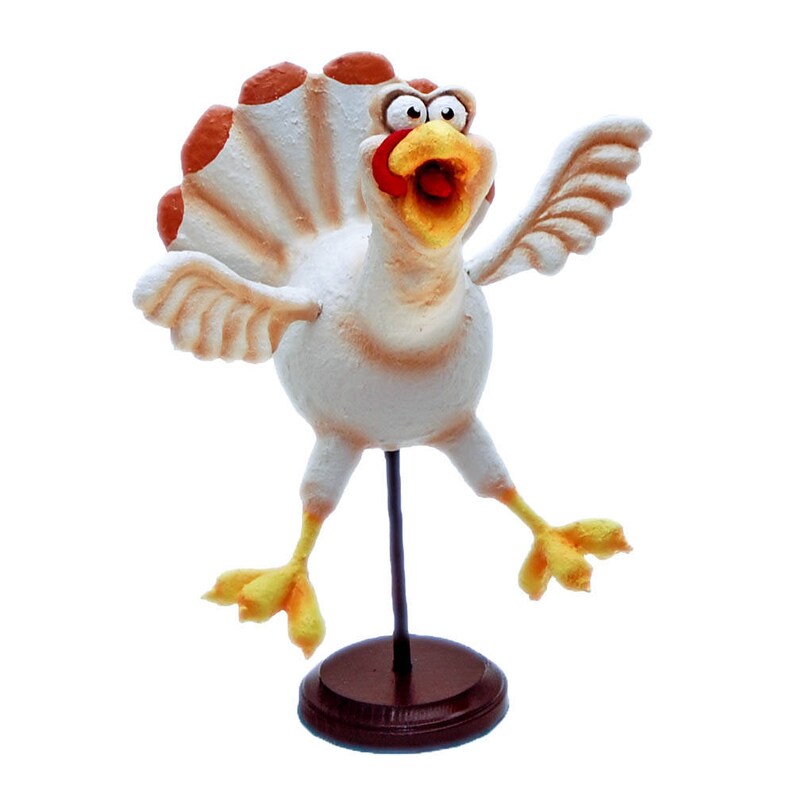 May include: A white turkey figurine with orange and brown feathers, yellow feet, and a red and yellow beak. The turkey is standing on a brown wooden base.
