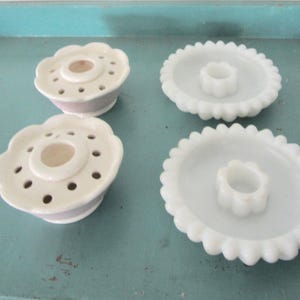 May include: Four vintage flower frogs. Two are white ceramic with a scalloped edge and a central flower-shaped holder. The other two are white ceramic bowls with a flower-shaped center and holes for flower stems.