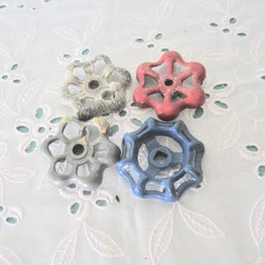 May include: Four vintage valve handles in various colors: gray, red, and blue. Each handle has a star-shaped design with a central hole. The handles are arranged on a white, floral-patterned fabric.