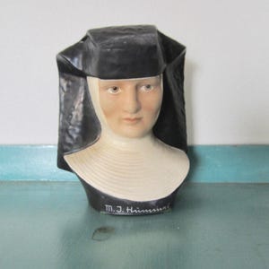 May include: A ceramic bust of a nun, featuring a black habit and a white wimple. The face is painted with realistic features, and the bust is signed "M.J. Hummel" at the base. The bust is set against a light background.