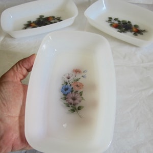 May include: A set of three white, rectangular serving dishes. One dish is held by a hand. Each dish features a floral or fruit design in the center. The dishes are made of a translucent material.