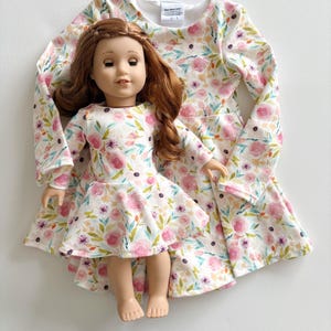 May include: A doll and matching dress set. The doll has auburn hair and is wearing a white dress with a pink floral pattern. The dress has long sleeves and a flared skirt. A matching dress is behind the doll.