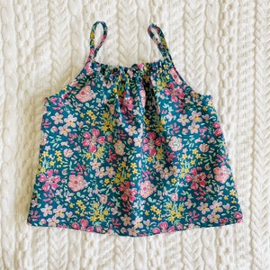May include: A sleeveless, teal blue tank top with a floral print featuring pink, yellow, and white flowers. The top has a gathered neckline and thin straps.