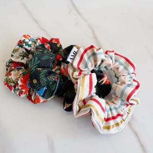May include: A collection of five fabric scrunchies in various patterns. One features a white background with red and green holly and poinsettia designs. Another has a black background with green and gold botanical elements. The third has red, white, and gold stripes.