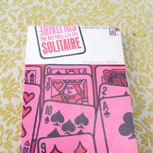 May include: Vintage book cover for "The Key Tool to Solitaire" by Douglas Brown. The cover features a pink and red design with playing card illustrations. The text "SOLITAIRE" is in a pink box. The book is a "New Imperial Book".