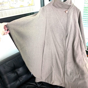 May include: A taupe cape coat with a high collar and a single button closure. The coat has wide sleeves and a flowing silhouette, suitable for layering. The fabric appears to be a soft wool blend, offering warmth and style.