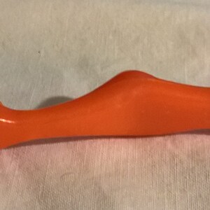 May include: An orange-colored tool with a metal tip. The tool has a curved, ergonomic handle and a pointed end. The tool is designed for a specific purpose, likely related to crafts or DIY projects.