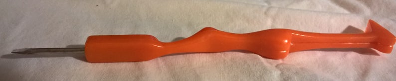 May include: An orange-colored tool with a metal tip. The tool has a curved, ergonomic handle and a pointed end. The tool is designed for a specific purpose, likely related to crafts or DIY projects.