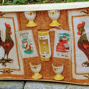 May include: A set of four yellow and white ceramic egg cups with sunflower designs, packaged with two white towels featuring rooster illustrations and the text "Country Style".