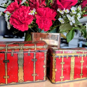 May include: Two antique red metal storage boxes with gold trim and accents. The boxes have a trunk-like design with decorative clasps and straps. A bouquet of red carnations and white flowers is in the background.