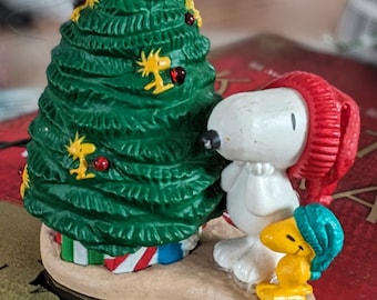 May include: A white ceramic figurine of Snoopy wearing a red hat standing next to a green Christmas tree with red and yellow ornaments. The figurine and tree are on a tan base.