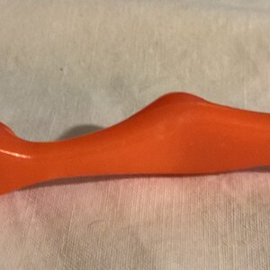 May include: An orange-colored awl tool with a metal tip. The awl has a curved handle for grip and a pointed end for piercing materials. The tool is designed for tasks such as sewing, leatherwork, or crafting.