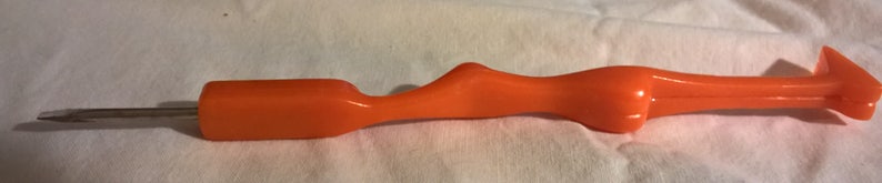 May include: An orange-colored awl tool with a metal tip. The awl has a curved handle for grip and a pointed end for piercing materials. The tool is designed for tasks such as sewing, leatherwork, or crafting.