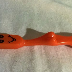 May include: An orange plastic cocktail fork with a stainless steel prong. The handle features a cartoon face with the word "cee" written above it. The fork is designed with a curved body and a flat end.