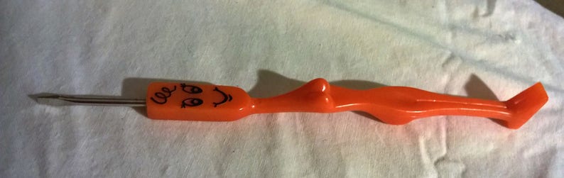 May include: An orange plastic cocktail fork with a stainless steel prong. The handle features a cartoon face with the word "cee" written above it. The fork is designed with a curved body and a flat end.